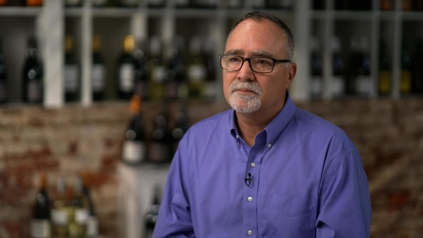 Greg Jones on 60 Minutes sitting in wine shop with bottles blurred in the background.